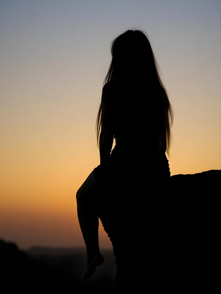 Silhouette: a girl sits on a cliff at dawn, dangling her legs. Everything is in black silhouette. Long hair falls down her back.