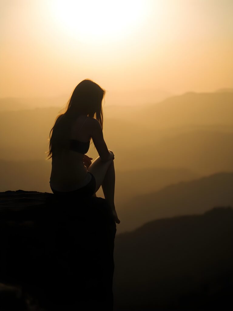Silhouette: a girl sits on a cliff at dawn, dangling her legs. Everything is in black silhouette. Long hair falls down her back.