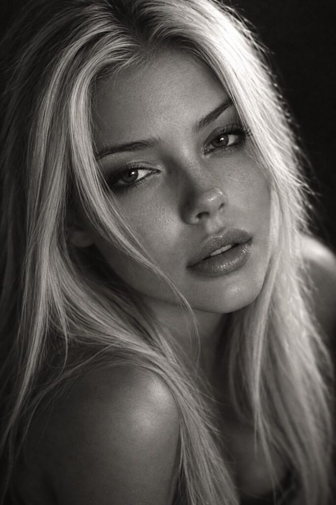 Sensual, dreamy black-and-white fine-art portrait photograph of a beautiful young woman in her mid-twenties with long, straight, very light platinum-blonde hair falling softly around her face and shoulders. Dark, deep, expressive eyes with a soft, alluring, half-lidded gaze looking into the camera; small cute upturned button nose; full soft lips slightly parted; flawless natural skin with realistic texture. Tight close-up focused on her face, head slightly tilted, a few strands of hair brushing her cheek, bare shoulders hinted at the bottom of frame. Perfect studio lighting: soft directional Rembrandt key light from the side, gentle sculpting shadows along cheekbone and jawline, subtle rim light glowing through the hair, catchlights in the eyes. Monochrome, rich tonal range, deep velvety blacks and luminous silvery highlights, fine film grain, shallow depth of field with creamy dark background, 85mm f/1.4 lens, editorial cinematic mood, seductive yet elegant and tasteful, dreamy soft glow.